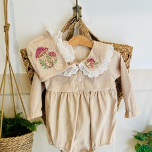 Pode incluir: Um macacão de bebê de veludo cotelê bege com mangas compridas e gola de renda branca. O macacão tem um bordado de cogumelos na frente e um chapéu combinando bordado com cogumelos com acabamento de renda branca.