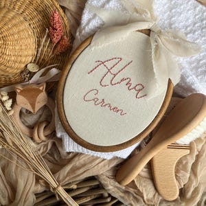 May include: An oval embroidery hoop with the name "Alma Carmen" stitched in red thread. A wooden baby brush and a wooden fox-shaped teether are also visible. The items are arranged on a woven basket and soft fabric.