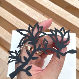 May include: A black paper cutout featuring a botanical design with two blooming flowers, a small bud, and several leaves on a single stem. The intricate silhouette is crafted from flat, matte black material with clean, sharp edges throughout the design.