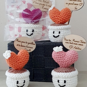 Peut inclure: Décorations faites à la main en forme de cœur au crochet, dans des pots blancs avec des visages souriants. Les cœurs sont dans des tons de rose et d'orange. Certains ont des étiquettes en bois avec du texte en français.