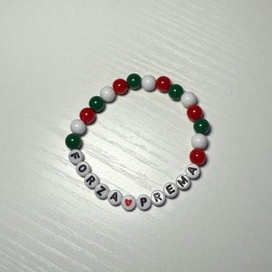 May include: A white beaded bracelet with red and green beads. The bracelet spells out "FORZA ♥ PREMA" in black letters.