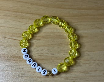 Puede incluir: Una pulsera de cuentas amarillas con el nombre "NORRIS" escrito con cuentas de letras blancas. Un pequeño dije de estrella dorada separa el nombre. La pulsera está hecha de cuentas amarillas facetadas.