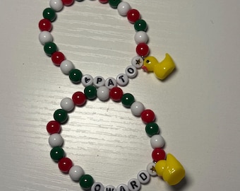 Pode incluir: Duas pulseiras de contas com contas vermelhas, brancas e verdes. As pulseiras soletram os nomes "PATTO" e "OWARD" com contas de letras pretas. Cada pulseira tem um amuleto de pato de borracha amarelo.