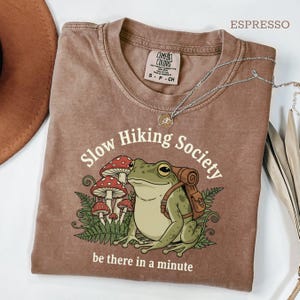May include: Espresso-colored t-shirt with a graphic of a frog wearing a backpack, surrounded by mushrooms and ferns. The text reads "Slow Hiking Society" and "be there in a minute". A silver necklace is draped across the shirt.