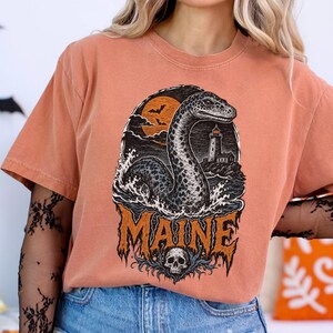 May include: A terracotta-colored short-sleeve t-shirt featuring a graphic print of a sea serpent rising from waves near a lighthouse under a moon with bats. The word MAINE is printed in large orange letters above a skull at the bottom of the design.