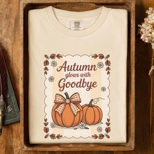 May include: Cream-colored t-shirt with a fall-themed graphic. The graphic features two orange pumpkins, one with a plaid bow, and the text "Autumn glows with Goodbye". The design is framed with floral and leaf elements.