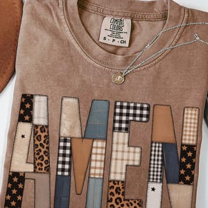 May include: A brown Comfort Colors t-shirt with the word "AMEN" appliqued across the front in a patchwork design. The letters feature various patterns, including stars, leopard print, and plaid. A silver necklace with a pendant is draped on the shirt.