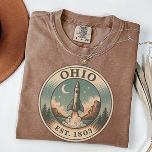 K&ouml;nnte beinhalten: Ein braunes Comfort Colors T-Shirt mit einem kreisf&ouml;rmigen Grafikdesign mit der Aufschrift "OHIO" und "EST. 1803". Die Grafik zeigt einen Raketenstart, einen Mond und eine Landschaft. Eine silberne Halskette liegt auf dem Shirt. Gr&ouml;&szlig;e S-CH.