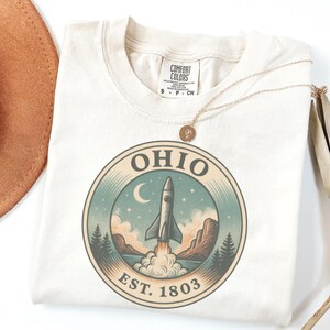 K&ouml;nnte beinhalten: Cremefarbenes T-Shirt mit einem kreisf&ouml;rmigen Grafikdesign, das eine Rakete zeigt, die &uuml;ber einer Landschaft mit Bergen, B&auml;umen und einem Halbmond startet. Das Wort "OHIO" und "EST. 1803" sind aufgedruckt. Eine goldene Halskette liegt auf dem Shirt.