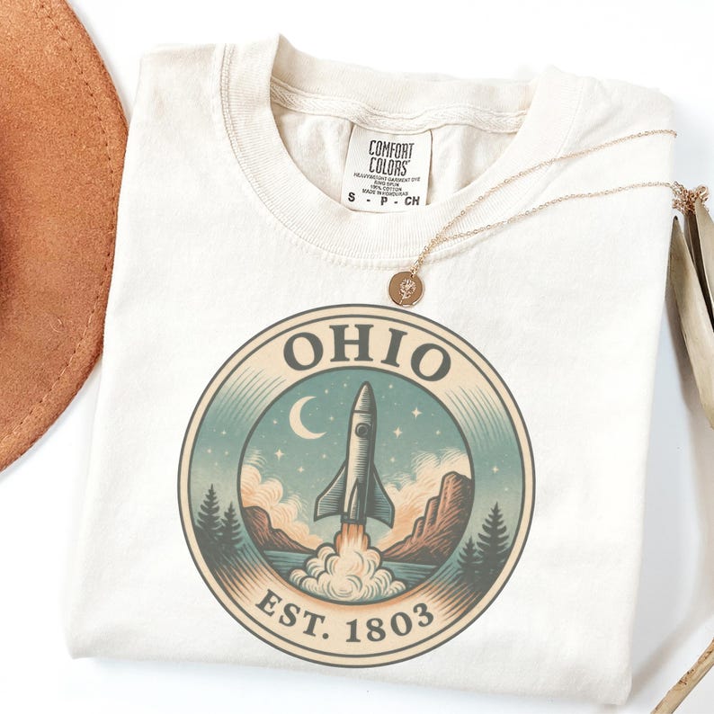 K&ouml;nnte beinhalten: Cremefarbenes T-Shirt mit einem kreisf&ouml;rmigen Grafikdesign, das eine Rakete zeigt, die &uuml;ber einer Landschaft mit Bergen, B&auml;umen und einem Halbmond startet. Das Wort "OHIO" und "EST. 1803" sind aufgedruckt. Eine goldene Halskette liegt auf dem Shirt.