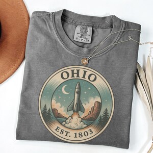 K&ouml;nnte beinhalten: Ein graues Comfort Colors T-Shirt mit einem kreisf&ouml;rmigen Grafikdesign, das eine Rakete zeigt, die von einer Landschaft startet, mit dem Text "OHIO EST. 1803". Eine silberne Halskette mit Anh&auml;nger liegt auf dem Shirt.