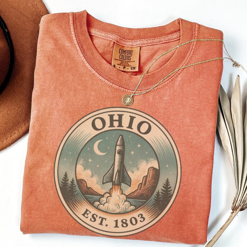 K&ouml;nnte beinhalten: Ein korallenfarbenes T-Shirt mit einem kreisf&ouml;rmigen Aufdruck, der eine Rakete beim Start in den Weltraum, einen Halbmond und das Wort "OHIO" &uuml;ber "EST. 1803" zeigt. Eine goldene Halskette mit Anh&auml;nger liegt auf dem Shirt.