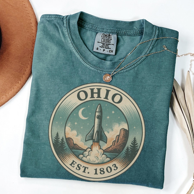 May include: Teal-colored t-shirt with a circular graphic featuring a rocket launching from a landscape with mountains, trees, and a crescent moon. The word "OHIO" is at the top, with "EST. 1803" at the bottom. A gold necklace is draped across the shirt.