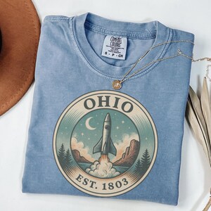 K&ouml;nnte beinhalten: Hellblaues T-Shirt mit einem runden Grafikdesign, das eine Rakete zeigt, die von einer Landschaft startet, mit dem Wort "OHIO" und "EST. 1803". Das Shirt hat einen Rundhalsausschnitt und kurze &Auml;rmel. Eine goldene Halskette liegt &uuml;ber dem Shirt.