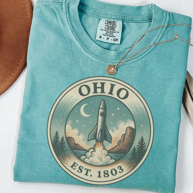 K&ouml;nnte beinhalten: Ein t&uuml;rkisfarbenes T-Shirt mit einem kreisf&ouml;rmigen Aufdruck, der eine Rakete zeigt, die von einer Landschaft mit Bergen, B&auml;umen und einem Halbmond startet. Das Wort "OHIO" w&ouml;lbt sich &uuml;ber der Rakete, darunter "EST. 1803". Eine goldene Halskette liegt auf dem Shirt.