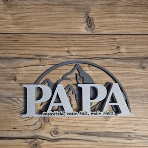 Può includere: Un cartello di legno grigio con la parola "PAPA" a grandi lettere, su uno sfondo di montagne. Sotto, la frase "meinHeld, mein Fels, mein Herz". Il cartello è posto su uno sfondo di legno.