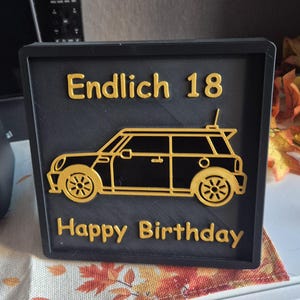 Könnte beinhalten: Schwarze quadratische Plakette mit goldfarbenen Buchstaben und einer goldenen Auto-Umrisslinie. Der Text lautet "Endlich 18" und "Happy Birthday". Das Auto hat ein detailliertes Design mit Rädern und einer Dachantenne. Die Plakette befindet sich auf einer gemusterten Oberfläche.