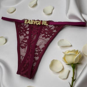 May include: Burgundy lace thong underwear with the word "BABYGIRL" in gold letters across the waistband. The underwear is displayed on a white satin surface, with white rose petals and a white rose in the background.