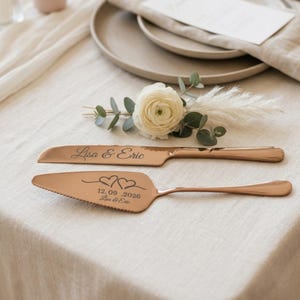 Puede incluir: Juego de cuchillo y servidor de pastel en oro rosa. El cuchillo está grabado con "Lisa & Eric", y el servidor tiene dos corazones y la fecha "12.09.2026". Un arreglo floral con una flor blanca y vegetación está en el fondo.