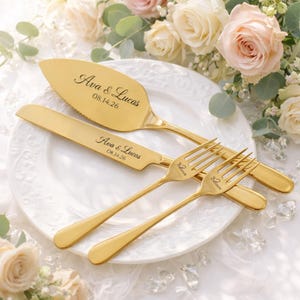 Peut inclure: Un ensemble doré pour servir le gâteau de mariage, comprenant une pelle, un couteau et deux fourchettes, gravés des noms "Ava & Lucas" et de la date "08.14.26". L'ensemble est présenté sur une assiette blanche.
