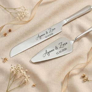 May include: A silver cake knife and server set. The knife is long and slender, while the server has a triangular blade with a serrated edge. Both are engraved with the names "Ayaan & Zara" and the date "12.10.2026".