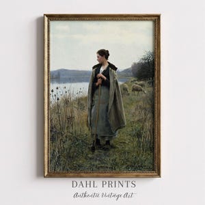 May include: Framed vintage print featuring a woman in a long cloak holding a staff, standing near a body of water. Sheep graze in the background. The print is in a gold-colored frame. The text "DAHL PRINTS Authentic Vintage Art" is at the bottom.