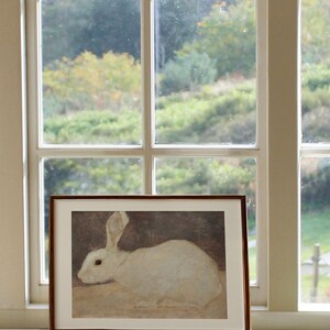 Puede incluir: Una impresi&oacute;n enmarcada de un conejo blanco, colocada en un alf&eacute;izar. La obra de arte se presenta sobre un fondo apagado, con una ventana y una vista de &aacute;rboles. El marco es marr&oacute;n y la impresi&oacute;n est&aacute; etiquetada como "DAHL PRINTS Authentic Vintage Art."