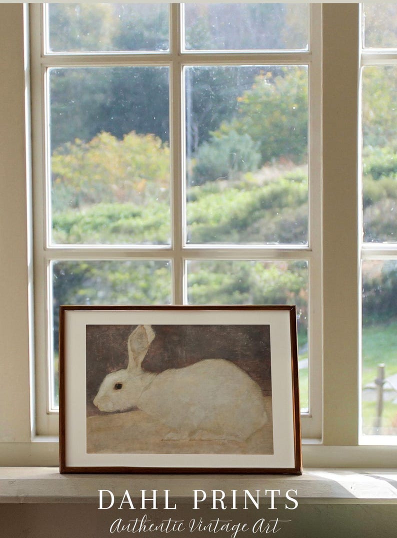 Puede incluir: Una impresi&oacute;n enmarcada de un conejo blanco, colocada en un alf&eacute;izar. La obra de arte se presenta sobre un fondo apagado, con una ventana y una vista de &aacute;rboles. El marco es marr&oacute;n y la impresi&oacute;n est&aacute; etiquetada como "DAHL PRINTS Authentic Vintage Art."