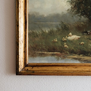 May include: An oil painting in a decorative gold frame depicting a mother duck and several ducklings resting in tall green grass near a calm body of water. The scene features muted natural tones, soft lighting, and a cloudy sky in the background.