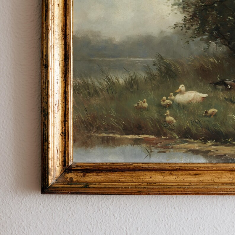 May include: An oil painting in a decorative gold frame depicting a mother duck and several ducklings resting in tall green grass near a calm body of water. The scene features muted natural tones, soft lighting, and a cloudy sky in the background.