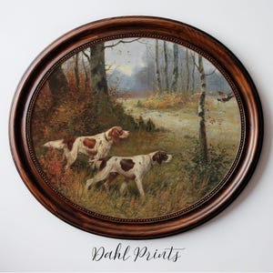 Puede incluir: Una impresión enmarcada ovalada que presenta dos perros de caza en una escena forestal. Los perros son blancos con manchas marrones. El marco es de madera marrón oscuro. La impresión está firmada "Dahl Prints" en cursiva en la parte inferior.