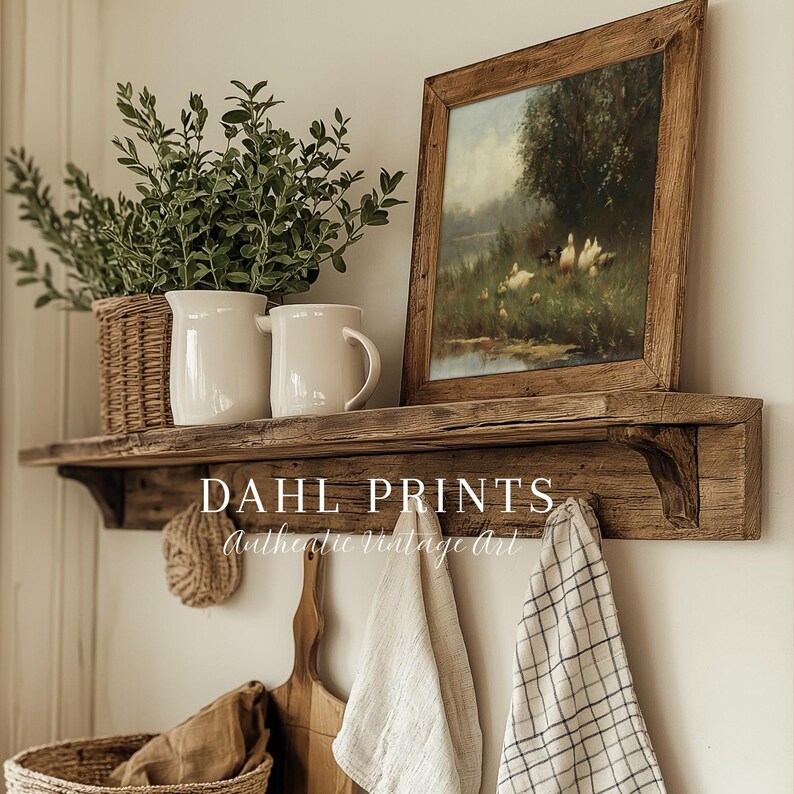 May include: A rustic wooden wall shelf featuring a framed landscape painting of ducks in a field, two white ceramic pitchers, a wicker basket with greenery, and hanging kitchen textiles. The text DAHL PRINTS Authentic Vintage Art is visible.