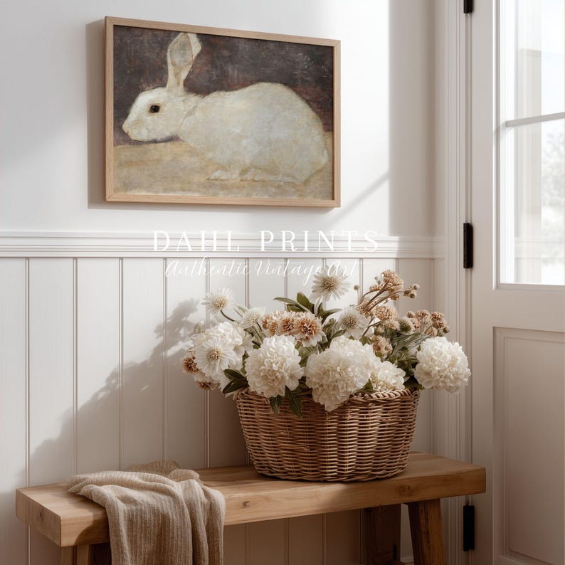 Puede incluir: Un cuadro enmarcado de un conejo blanco cuelga sobre un banco de madera. Una cesta tejida llena de flores blancas y beige est&aacute; sobre el banco. Una manta beige est&aacute; sobre el banco. La impresi&oacute;n est&aacute; en un marco de madera.