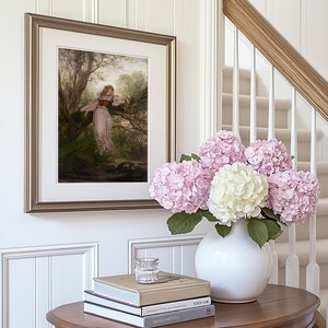 May include: A framed print featuring a painting of a figure in a light-colored dress and dark corset leaning against a tree in a forest. Beside it, a white ceramic vase holds a large bouquet of pink and white hydrangea flowers on a wooden table.