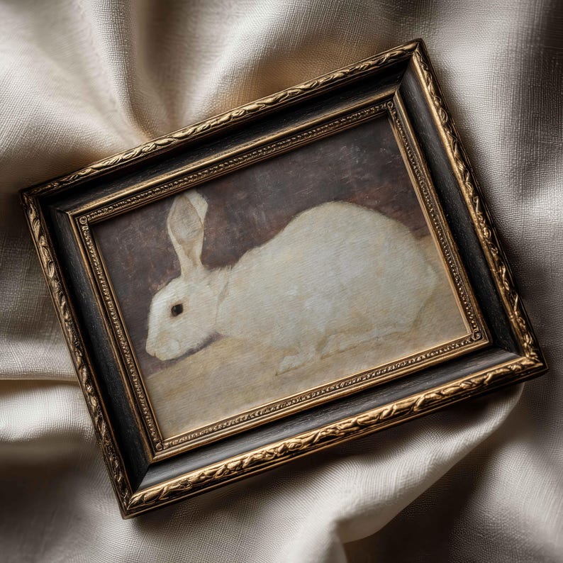Puede incluir: Pintura enmarcada de un conejo blanco en estilo vintage. La obra de arte est&aacute; enmarcada en un marco oscuro y ornamentado con detalles dorados. El conejo se representa en una pose relajada, con un fondo suave y neutro.