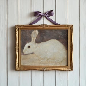Puede incluir: Pintura enmarcada de un conejo blanco con orejas largas, sobre un fondo oscuro. La pintura est&aacute; en un marco dorado adornado y est&aacute; rematada con un lazo de sat&eacute;n p&uacute;rpura. La obra de arte est&aacute; colgada en una pared de madera blanca.