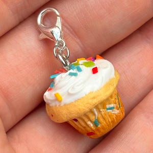 May include: A miniature cupcake charm with white frosting and colorful sprinkles. The charm has a silver-colored clasp for easy attachment to a bracelet or necklace. The cupcake has a yellow-brown paper cup.