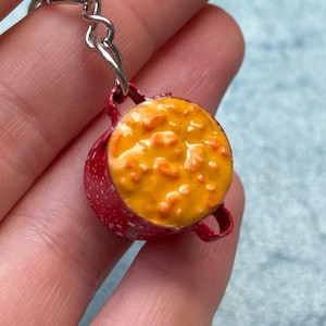 May include: A miniature red pot keychain filled with a yellow and orange stew. The pot has small handles and a silver chain. The stew appears to have a textured surface.