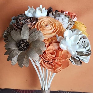 May include: A bouquet of handcrafted flowers in shades of orange, white, brown, and yellow. The bouquet features a large, daisy-like flower and is tied with twine. The stems are white.
