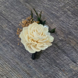 May include: A cream-colored sola wood flower boutonniere with a pearl accent and greenery.
