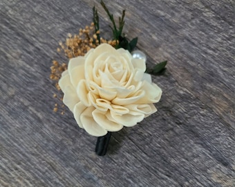 May include: A cream-colored sola wood flower boutonniere with a pearl accent and greenery.