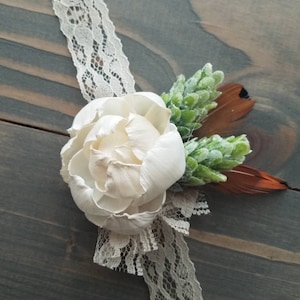 May include: A white flower with brown feathers and green foliage attached to a beige lace ribbon.
