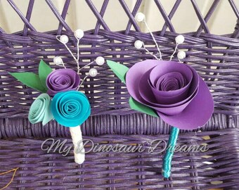Puede incluir: Dos boutonnières de flores de papel, una con una rosa morada y la otra con una rosa azul turquesa y azul claro. Ambas boutonnières tienen detalles de perlas y hojas verdes.