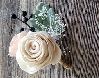 May include: A cream-colored sola wood rose with a light pink rose bud and sprigs of white baby's breath and green foliage. The bouquet is tied with twine.