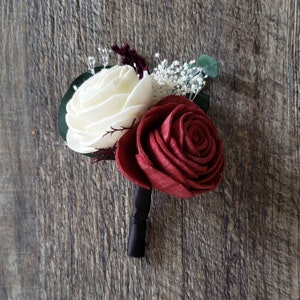 May include: A handcrafted boutonniere featuring a cream-colored and a burgundy rose, accented with small white flowers and greenery. The flowers are attached to a dark green base with a black ribbon-wrapped stem. Perfect for weddings or special events.