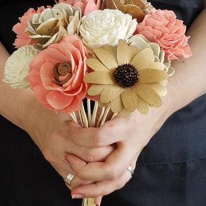 May include: A bouquet of sola wood flowers in shades of peach, white, and gold. The flowers are arranged in a hand-tied bouquet and have a rustic, natural look.
