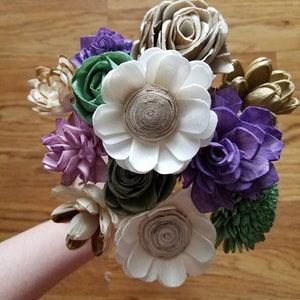 May include: A bouquet of wooden flowers in shades of white, green, purple, and gold. The flowers are arranged in a circular shape and have a rustic, natural look.