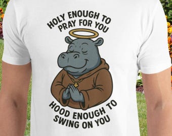 May include: White t-shirt featuring a cartoon hippo with a halo and a brown robe, hands clasped in prayer. The text reads "Holy Enough to Pray for You" and "Hood Enough to Swing on You".