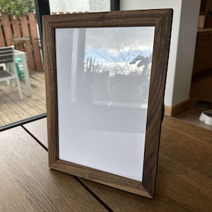 May include: A dark brown wooden picture frame with a white mount and a clear glass front. The frame is standing upright on a wooden table.