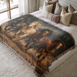 May include: A woven throw blanket featuring a painting of four horses running. The horses are depicted in shades of black, tan, and brown, with flowing manes and tails. The blanket has a fringed edge and is displayed on a bed.